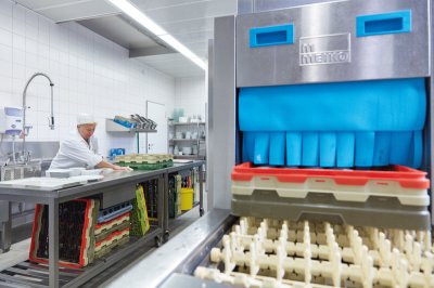 rechts: Geschirrspülmaschine einer Großküche, links im Hintergrund: Mitarbeiterin an einem Arbeitstisch
